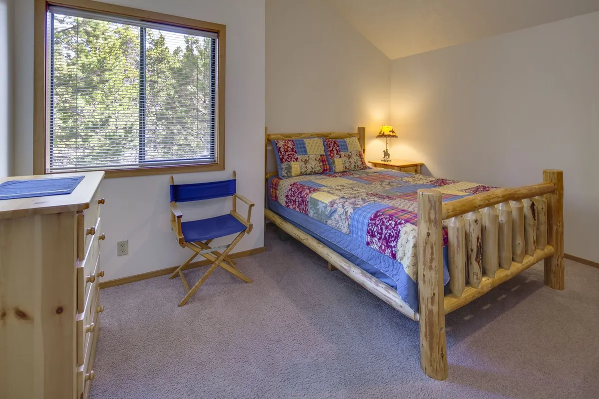 A tranquil bedroom with a log bed dressed in a colorful quilt, a blue reading chair, a lamp-lit nightstand, and a window framing the natural scenery outside. The inviting arrangement creates an intimate and restful setting, perfect for couples seeking a relaxing retreat.