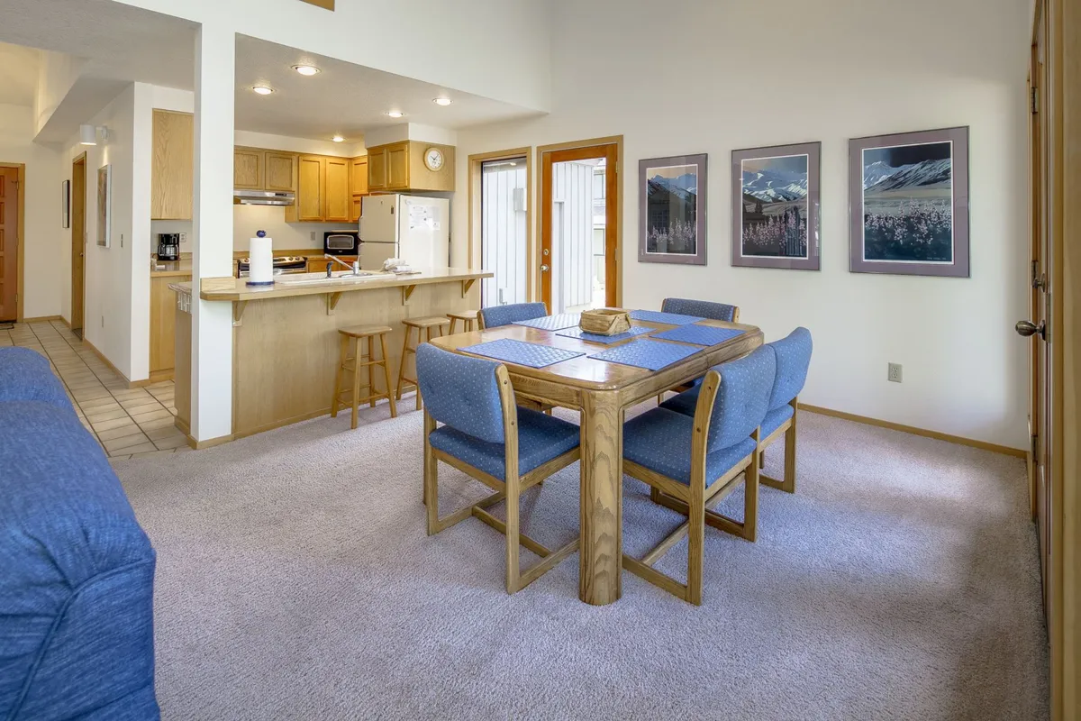 A bright, inviting dining area seamlessly connects to the kitchen. A generous dining table sits in the heart of the space, perfect for family meals or group celebrations. Sliding doors and framed photographs add warmth and a sense of openness, and the kitchen area is visible in the background.