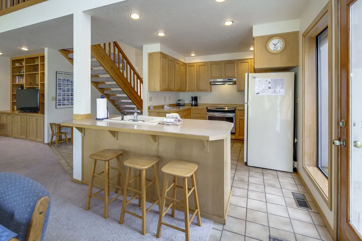 A view of the kitchen island surrounded by stools, with the staircase and cabinetry visible. The space is inviting for socializing, preparing group meals, or sharing a holiday feast, reflecting the section’s mention of BBQ dinners and communal dining.