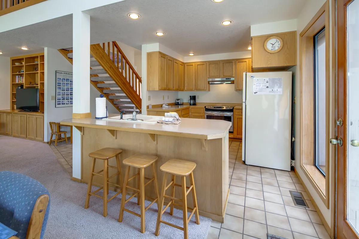 This image centers on the kitchen, where a prominent island, wooden cabinetry, and stainless steel appliances are featured. The space is open and easily accessible, designed for both cooking and socializing, with a staircase in view hinting at the home's multi-level layout.