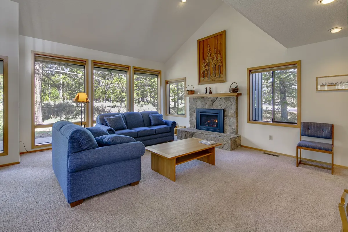 This image captures a warm and inviting living area, featuring blue sofas arranged around a fireplace. Large windows dominate one wall, flooding the space with natural light and framing views of the outdoors. The wooden coffee table and tasteful décor contribute to the cozy, communal atmosphere.