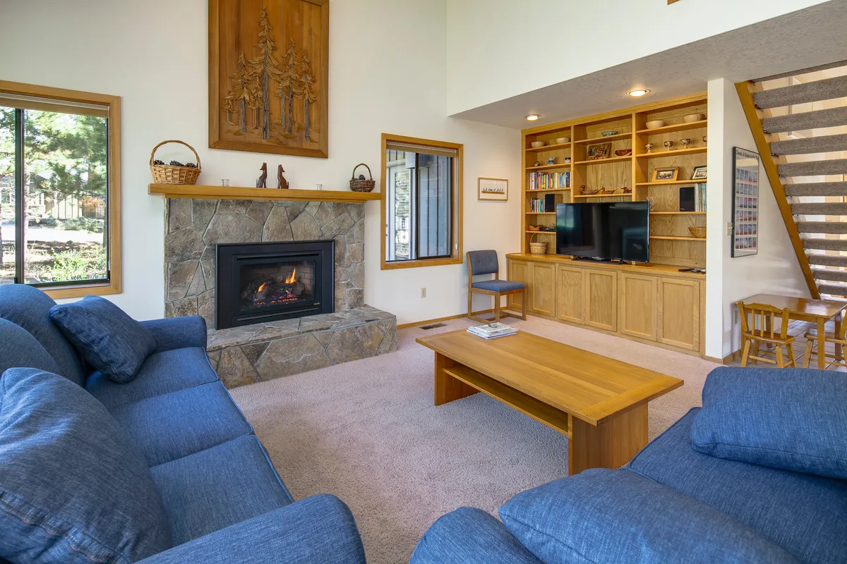 The image showcases a warm, inviting living room with a glowing fireplace as the centerpiece, surrounded by comfortable seating and wooden shelves. The decor is cozy and modern, perfect for relaxing or gathering with friends and family. The natural light from nearby windows highlights the spaciousness and intimacy of the setting.