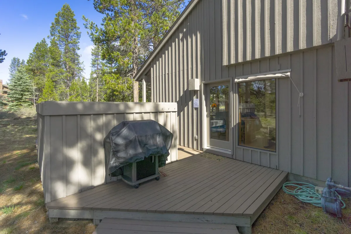 This image showcases a spacious deck area connected to the home, featuring a BBQ grill (covered with a tarp), large windows offering indoor-outdoor flow, and comfortable space for outdoor dining and barbecuing. The serene atmosphere is enhanced by the surrounding greenery, reflecting an inviting spot for meals and relaxation.